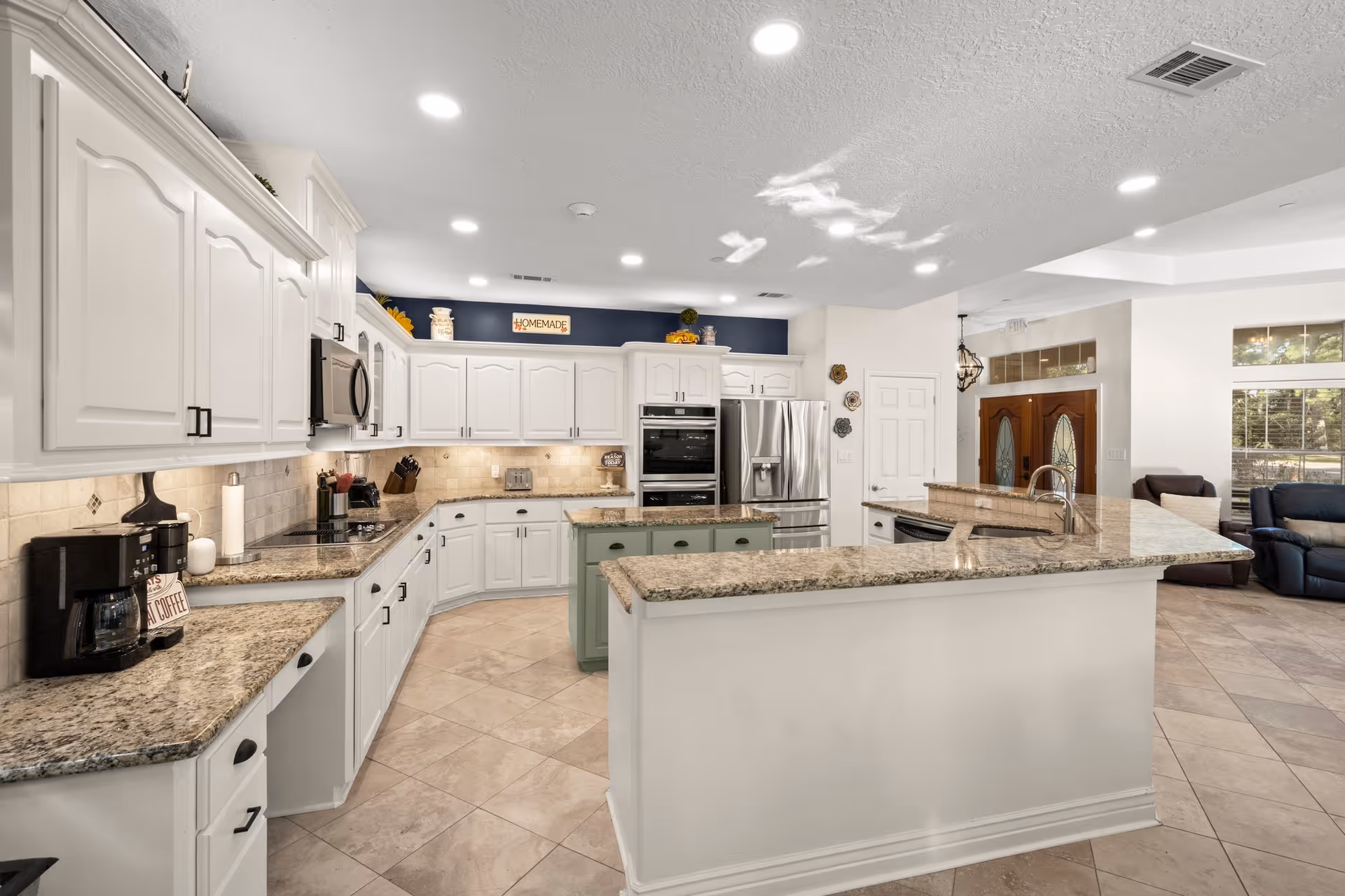 A spacious, modern kitchen with white cabinetry, granite countertops, and stainless steel appliances including a refrigerator and built-in oven. The kitchen features a large island with a sink and a smaller green island in the center. The floor is tiled, and the kitchen opens into a living area with large windows and a wooden front door visible in the background.