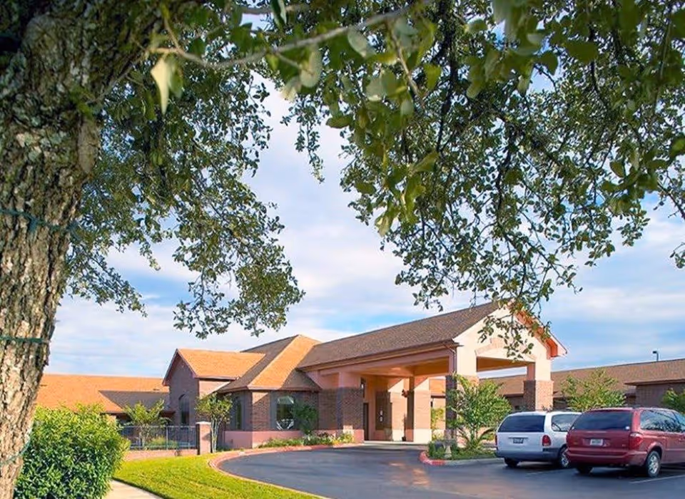 Exterior view of Kirkwood Manor Nursing and Rehab Center showing the front entrance with a covered driveway, surrounded by trees and parked cars in the parking lot under a partly cloudy sky.