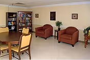 Community sitting area with a round wooden table and chairs, bookshelves, and two upholstered armchairs against a pale yellow wall.