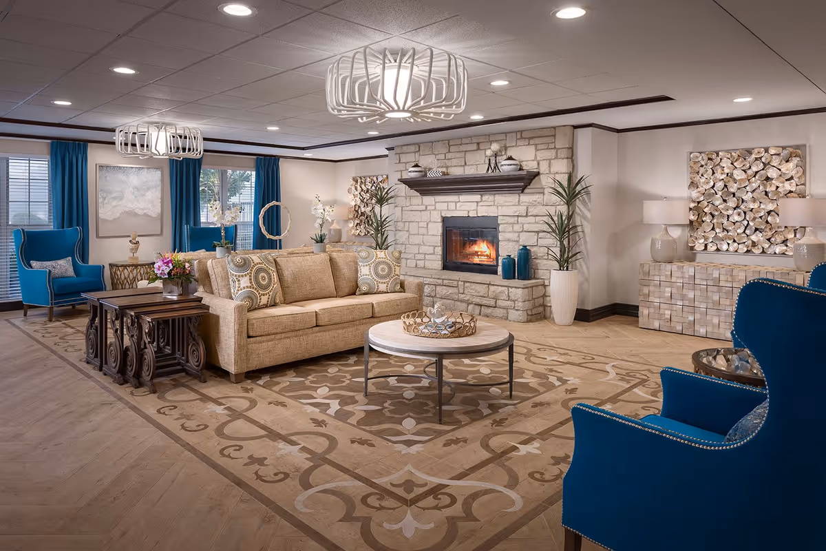 A spacious and elegantly decorated living room with a beige sofa, two blue armchairs, a round coffee table, and a stone fireplace with a fire burning. The room features a patterned area rug, modern light fixtures on the ceiling, blue curtains on the windows, and decorative plants and artwork on the walls.