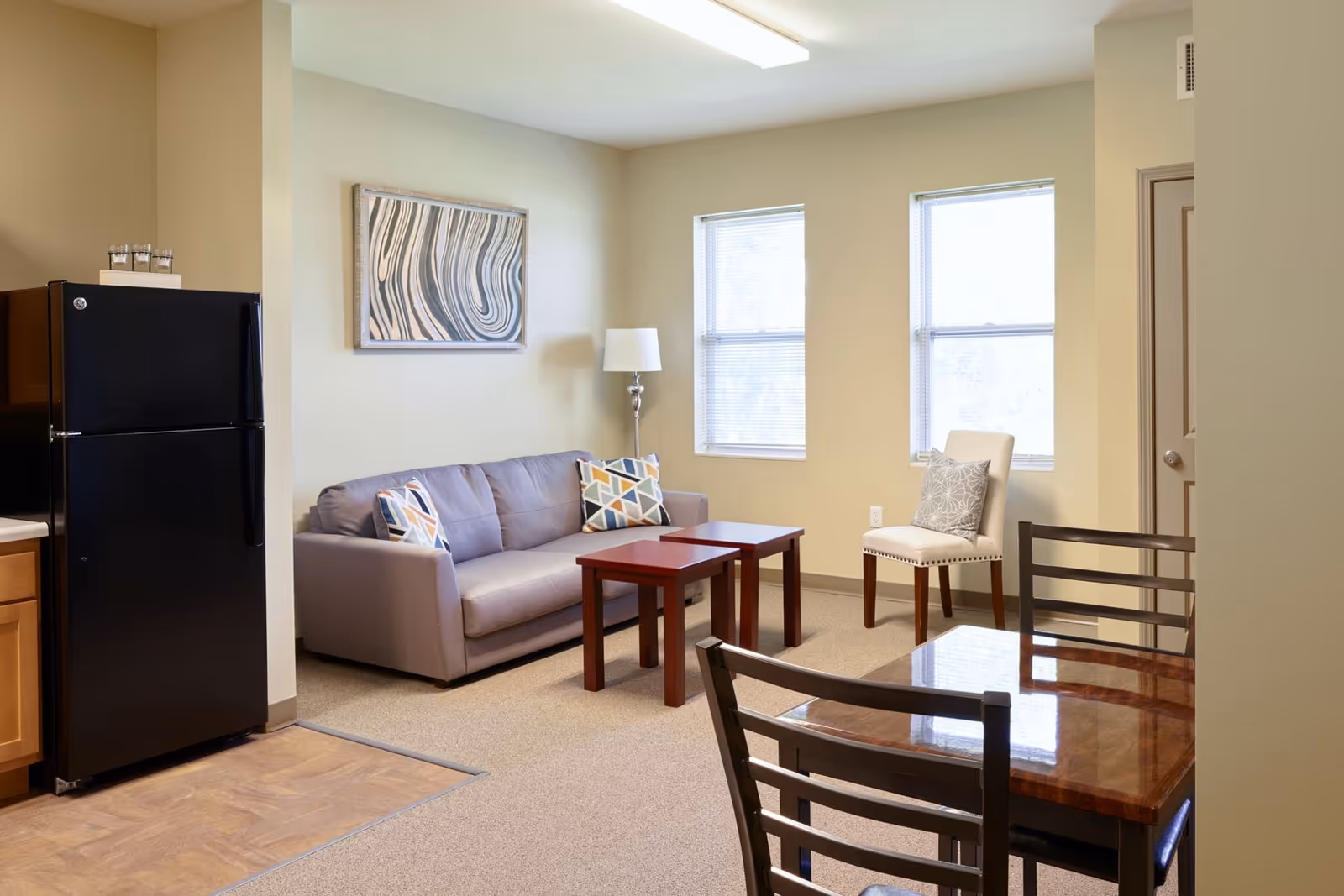 A cozy living area in a senior living facility featuring a gray sofa with patterned cushions, two small wooden coffee tables, a white upholstered chair with a decorative pillow, and two windows letting in natural light. To the left, there is a black refrigerator and part of a kitchen counter with wooden cabinets. A framed abstract artwork hangs on the wall above the sofa.