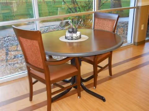 Round wooden dining table with two upholstered chairs beside a large window.