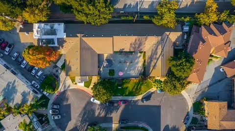 Aerial view of Silver Oaks Memory Care facility showing the building with a flat roof, surrounding trees, parked cars, and a small courtyard area with a pink umbrella.
