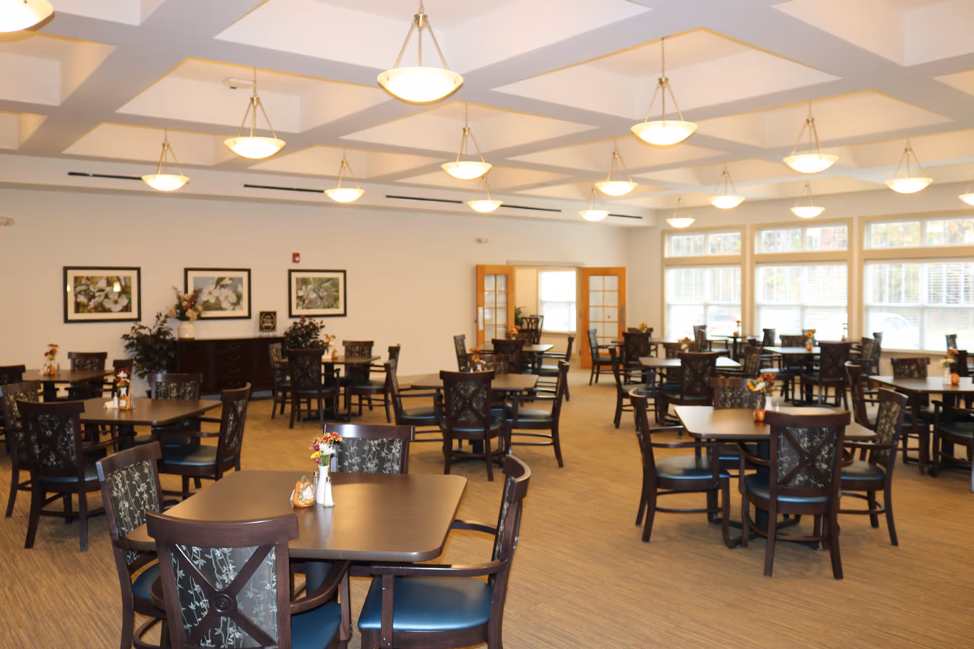 A spacious dining room with multiple dark wood tables and chairs arranged neatly. The room is well-lit with ceiling lights and large windows on one side allowing natural light to enter. There are framed floral pictures on the wall and small flower arrangements on each table.