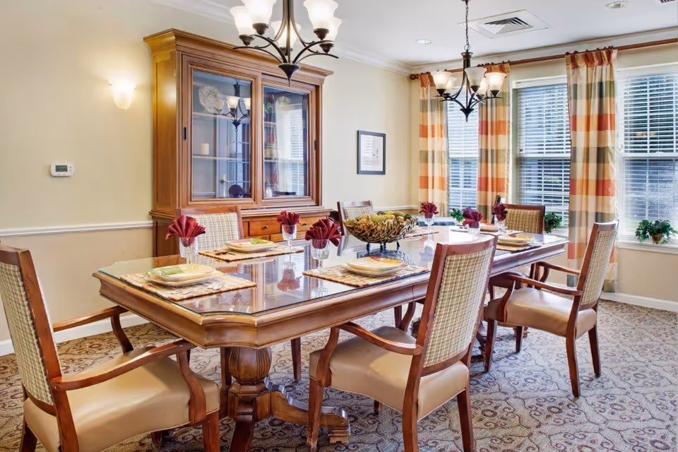 A well-lit dining room with a wooden dining table set for six people. The table has placemats, plates, glasses with folded red napkins, and a decorative centerpiece. Behind the table is a wooden china cabinet with glass doors. The room has large windows with striped curtains and patterned carpet flooring.