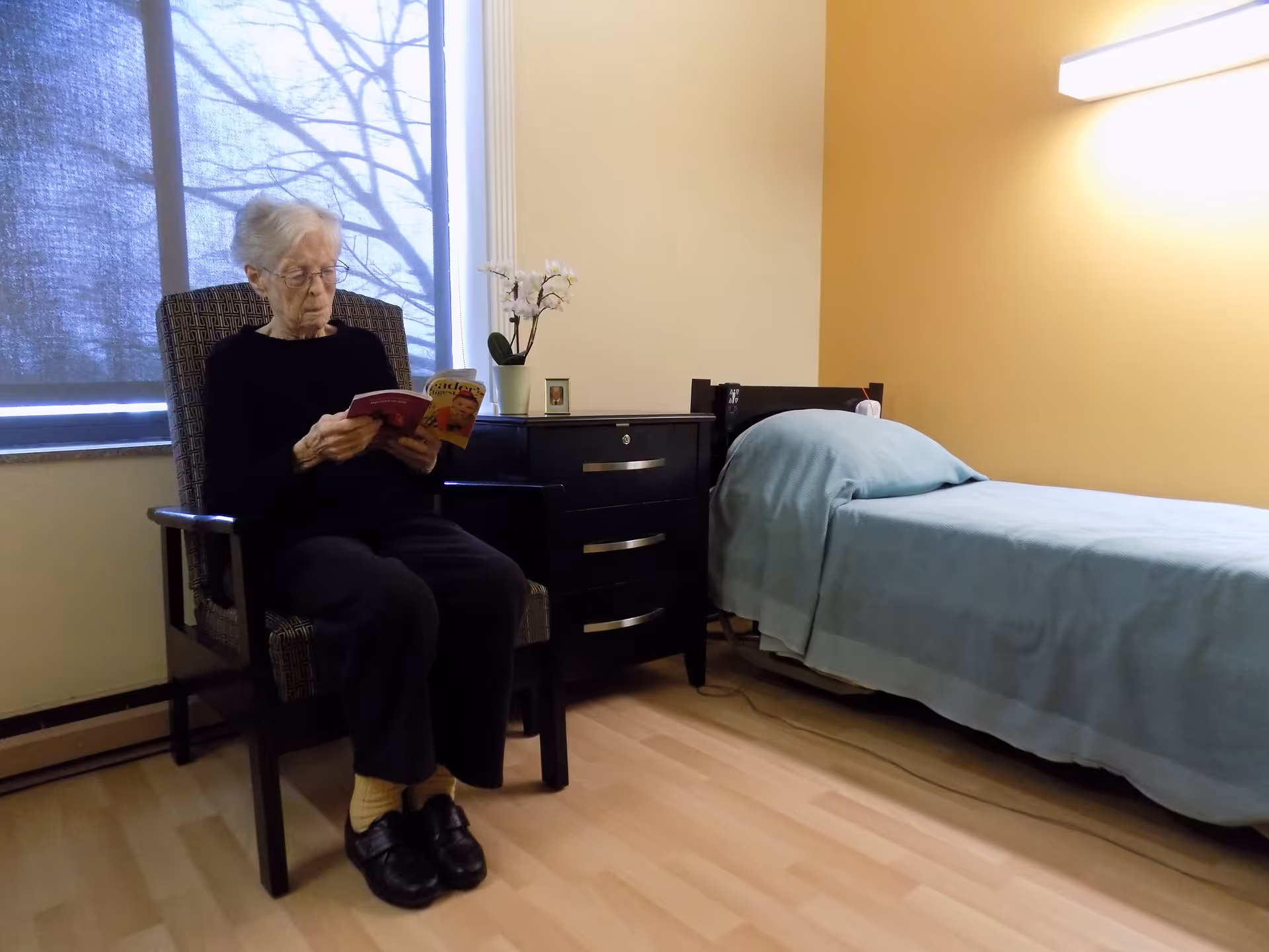 An elderly woman with white hair and glasses is sitting in a patterned armchair reading a book in a simple, well-lit room. Next to her is a black dresser with a small potted orchid and a framed photo on top. To the right is a single bed with a light blue blanket and pillow, against a yellow wall with a wall-mounted light above it. A window with a partially lowered blind is behind the woman.