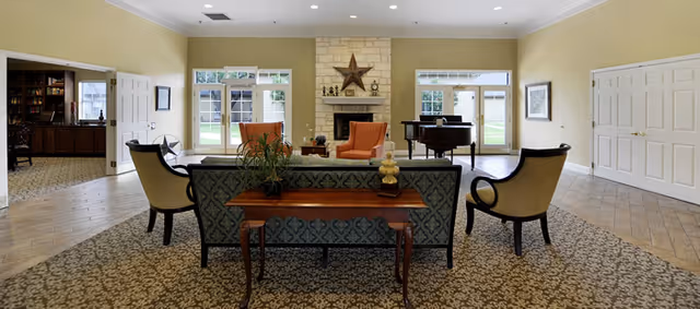 A spacious living room area in a senior living facility with a patterned carpet and tiled floor. The room features a stone fireplace with a star decoration above it, two orange armchairs, a grand piano near the windows, and a sofa with a wooden table behind it holding decorative items. Large windows and glass doors allow natural light to fill the room, and there are additional chairs arranged around the space.