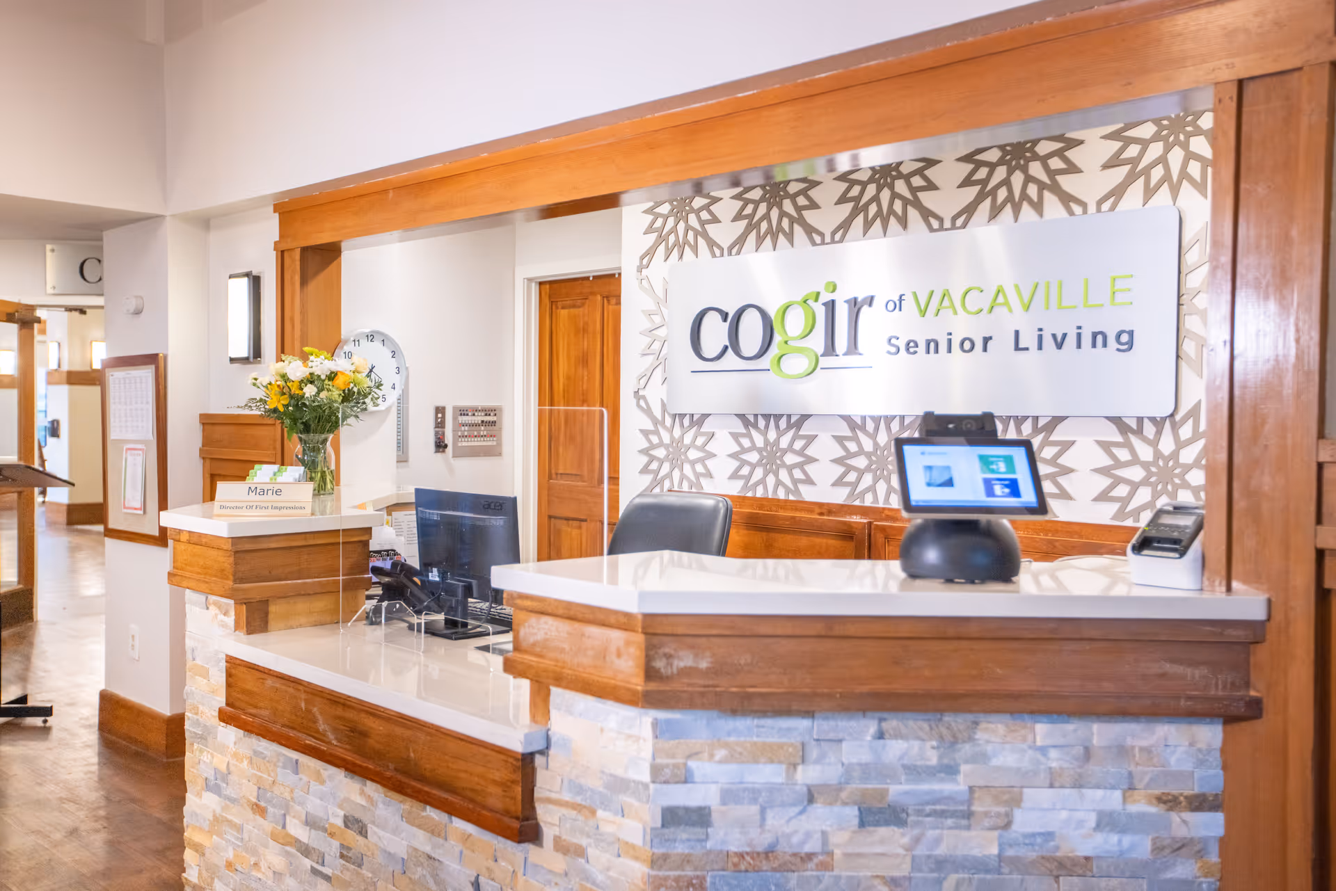 Reception desk area inside Cogir of Vacaville Senior Living facility with a computer, a vase of flowers, a nameplate reading 'Marie Director Of First Impressions', and a sign on the wall displaying the facility's name.
