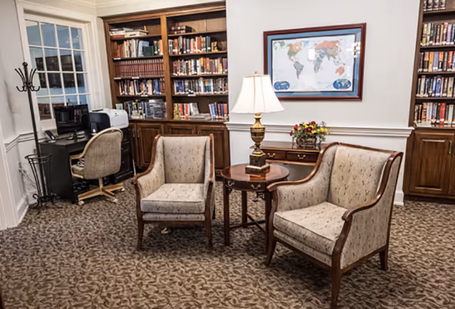 A cozy reading or sitting area in a senior living facility featuring two upholstered armchairs with wooden frames positioned around a small round wooden table with a decorative lamp and a flower arrangement. Behind the chairs are built-in wooden bookshelves filled with books, a framed world map on the wall, and a small desk with a computer and printer near a window.