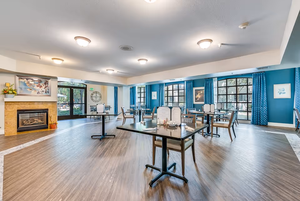 A bright and spacious dining room in a senior living facility with several tables and chairs arranged neatly. The room features large windows with blue curtains, a fireplace with a painting above it, and a wooden floor. Menus and condiments are placed on the tables.