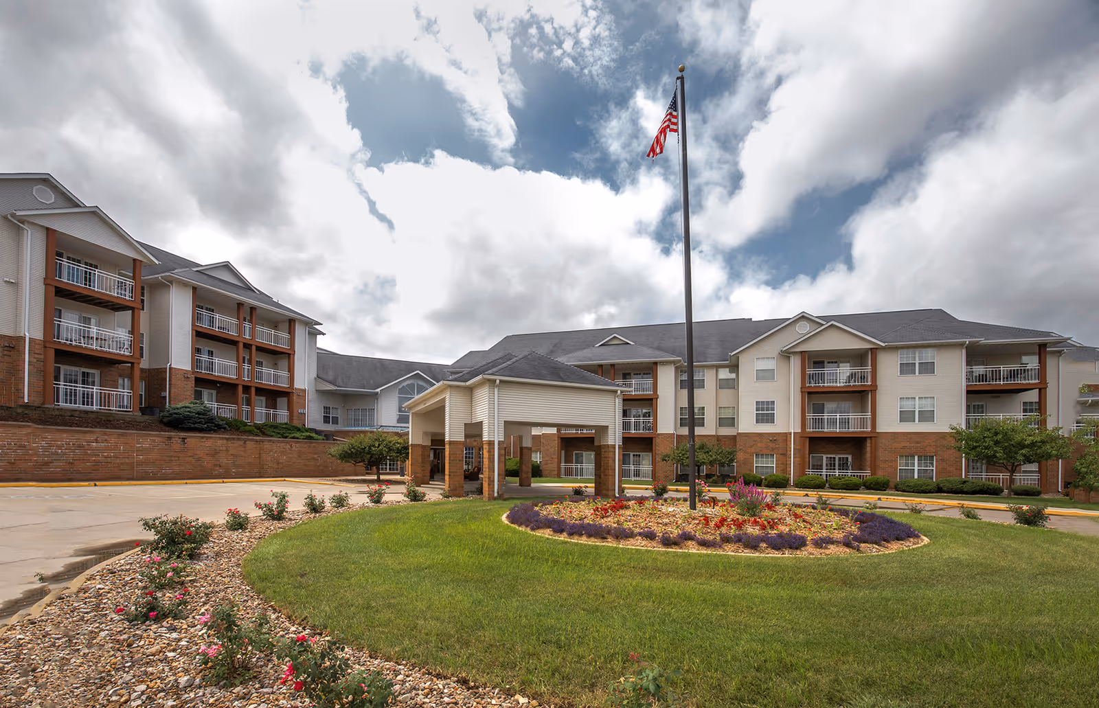 Front exterior of a multi-story senior living building with a covered entrance, landscaped circular driveway and an American flag.