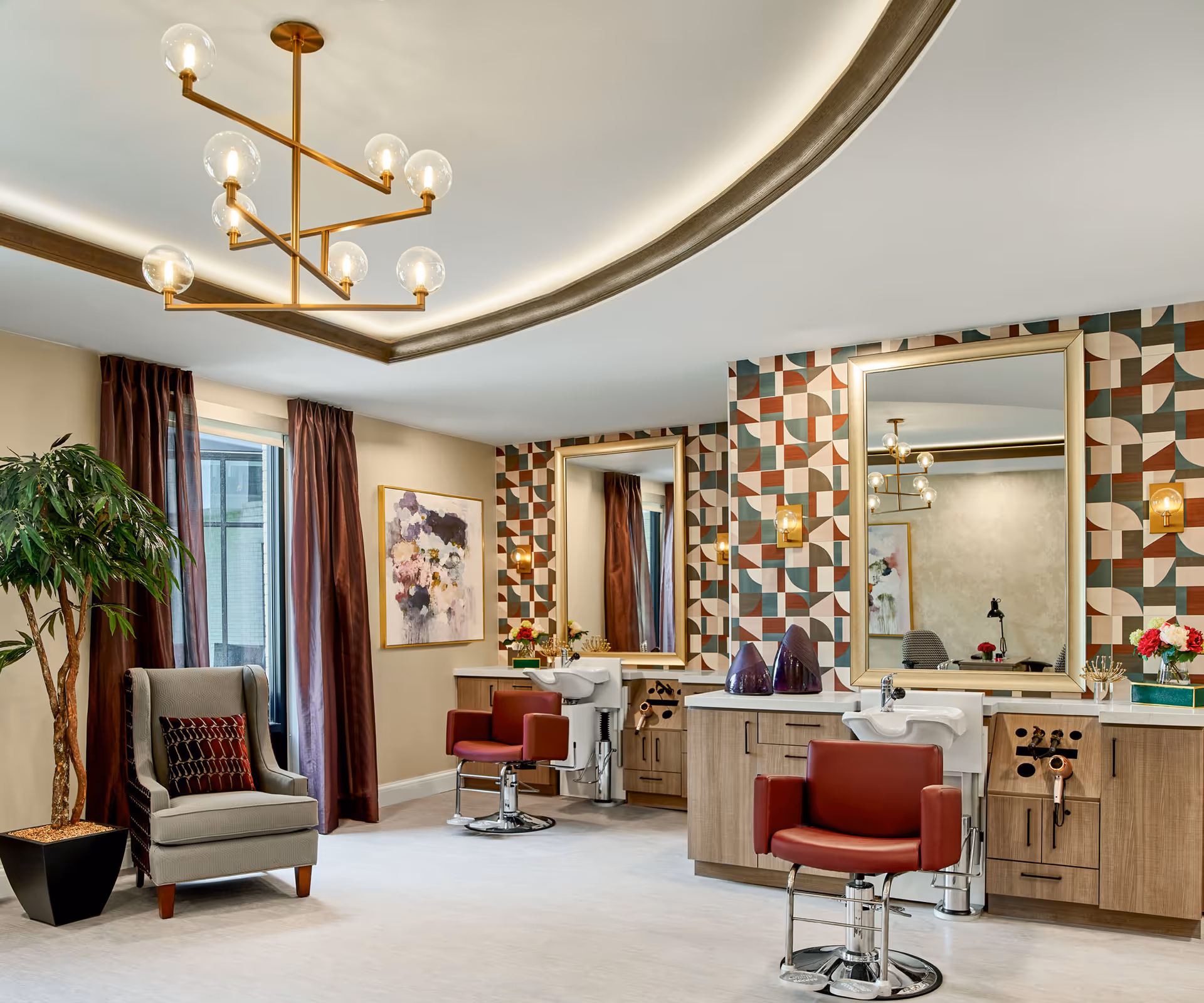 Interior of a senior living facility's salon area featuring two red salon chairs in front of large mirrors with geometric patterned wallpaper. There is a gray armchair with a decorative pillow next to a potted plant near a window with dark curtains. A modern chandelier with globe lights hangs from the ceiling.