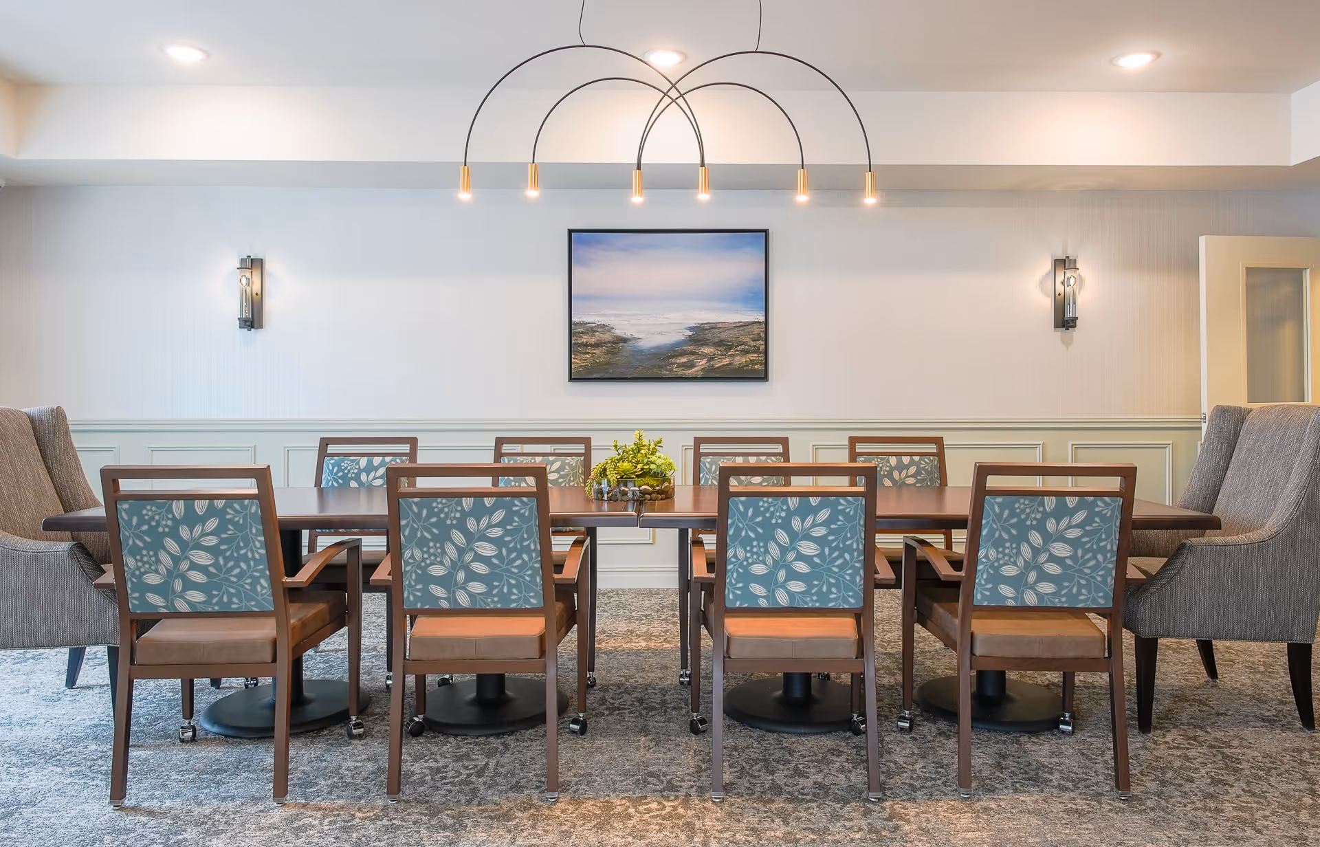 A well-lit dining room with a long wooden table surrounded by eight chairs, six with blue patterned backs and two large gray armchairs at the ends. A modern chandelier with five hanging lights is above the table. The walls are light-colored with wainscoting and two wall sconces. A framed landscape painting hangs on the wall behind the table.