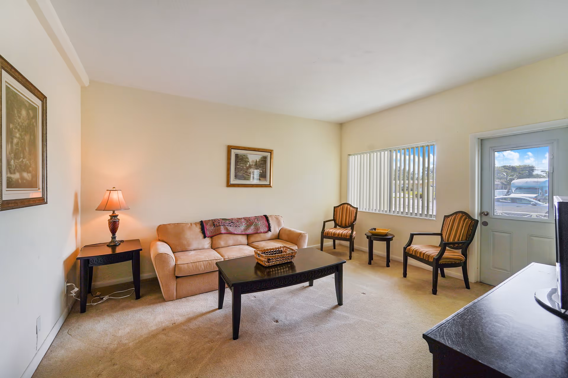 A cozy living room with a beige sofa, two striped armchairs, a dark wooden coffee table with a wicker basket, a side table with a lamp, framed artwork on the walls, a window with vertical blinds, and a door leading outside.