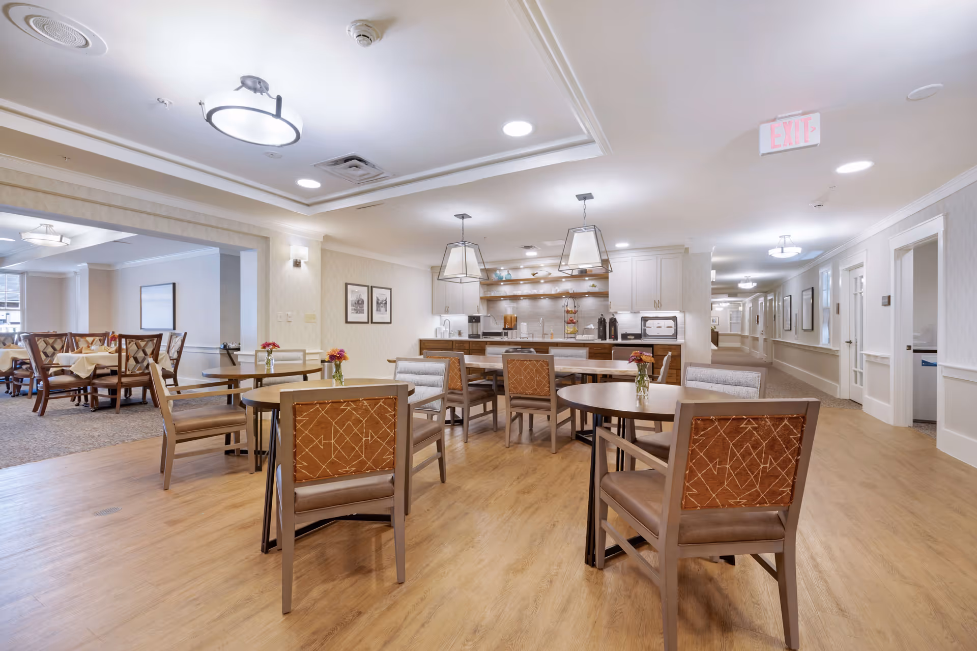 A bright and spacious dining area in a senior living facility with multiple round and rectangular tables surrounded by chairs. The room features wood flooring, modern pendant lights, and a kitchen area with cabinets, shelves, and appliances in the background. There are small flower vases on the tables and framed pictures on the walls.