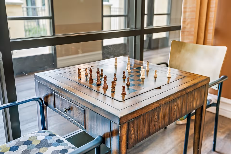A wooden chess table with a chess game in progress, positioned near large windows with natural light. Two chairs are placed on opposite sides of the table, one with a patterned cushion and the other with a plain seat. The room has a warm color scheme with orange walls and curtains.