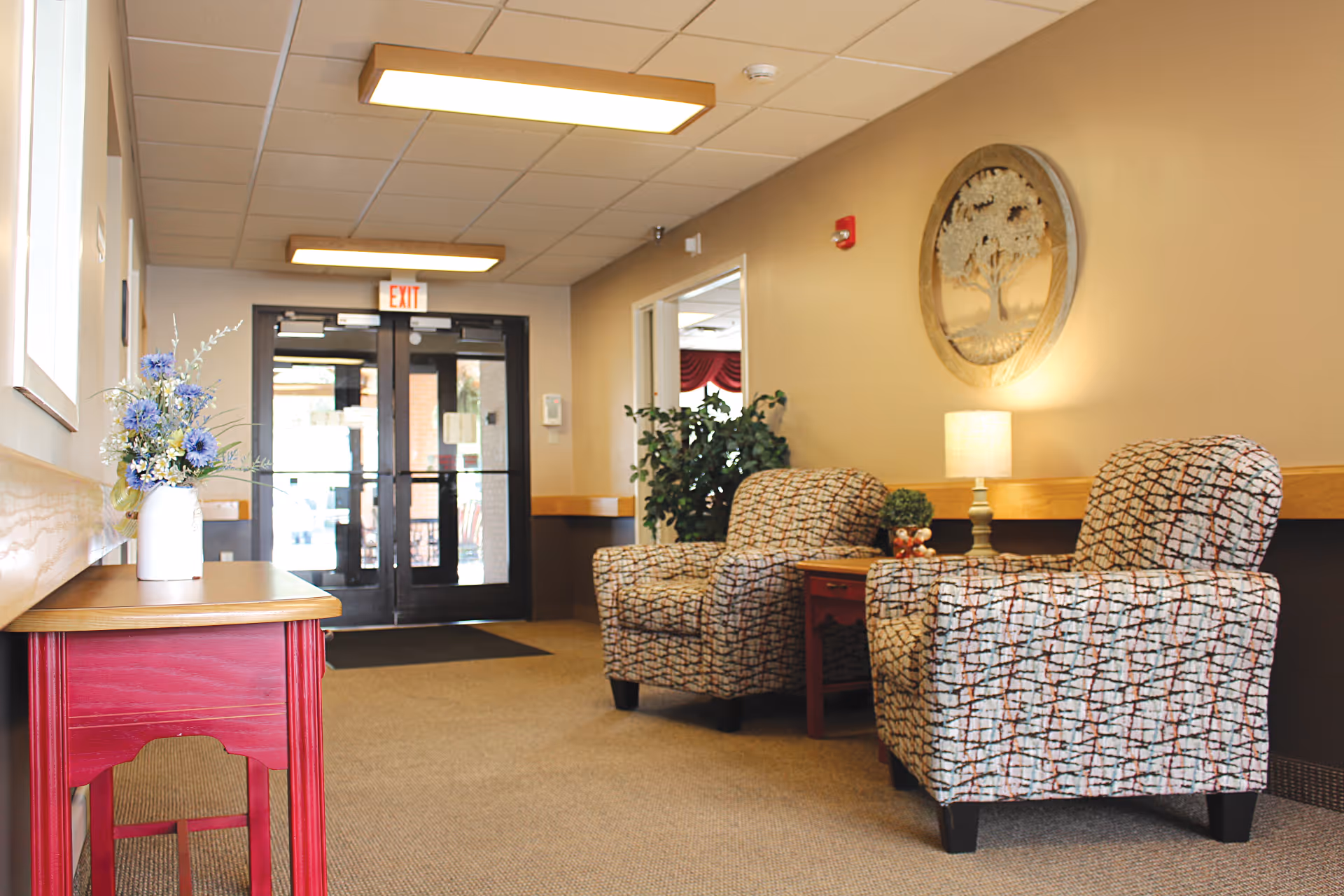 A cozy waiting area inside Mount Zion Health and Rehab Center featuring two patterned armchairs with a small wooden side table between them holding a lamp and a small plant. A red wooden table with a vase of flowers is on the left side near the window. The area has beige walls, carpeted floor, and a double glass door exit in the background with an exit sign above it. A decorative round wall art piece depicting a tree is mounted on the wall above the lamp.