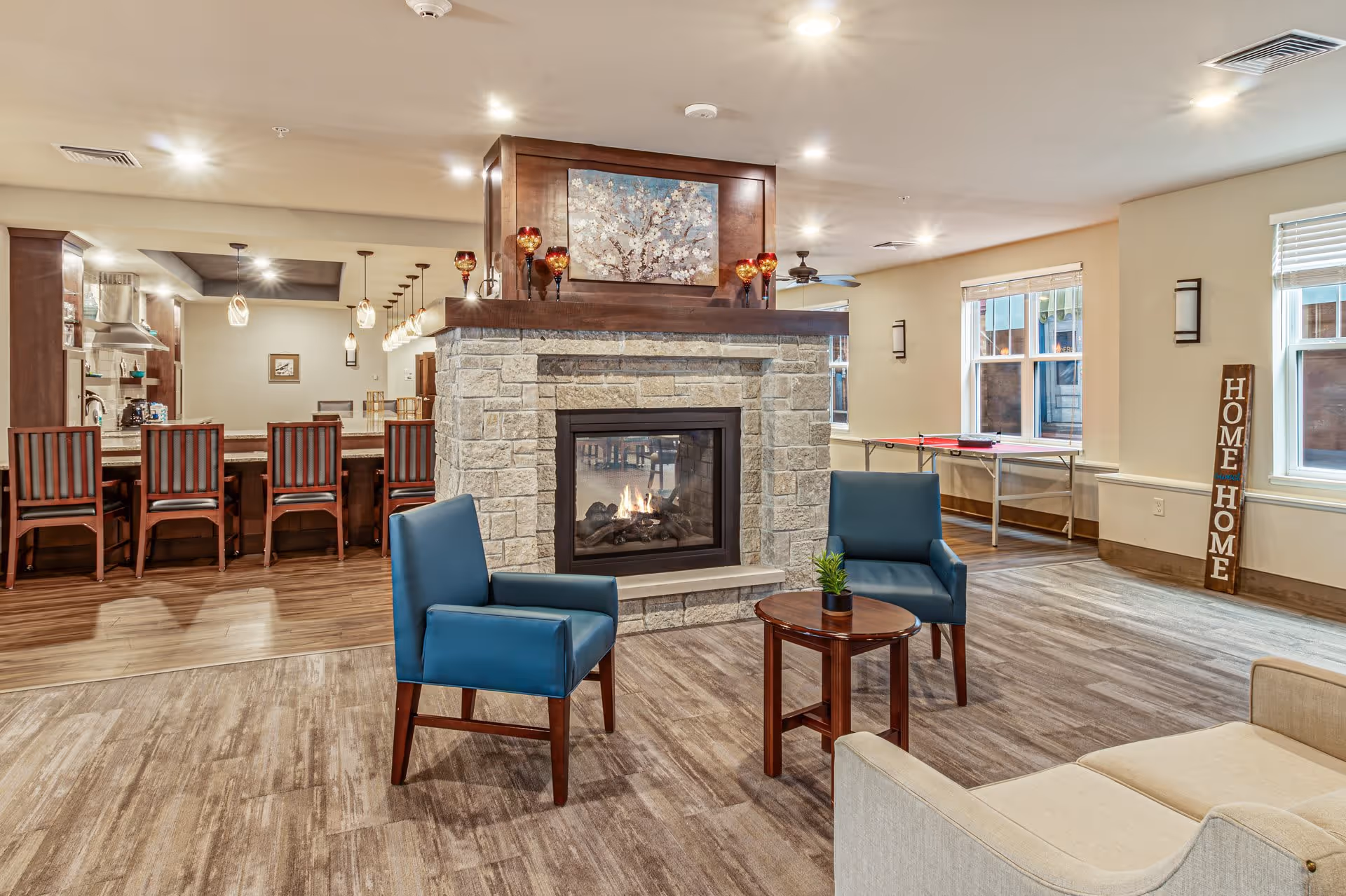 A cozy assisted living common area featuring a stone fireplace with a fire burning, two blue armchairs and a small round wooden table with a plant in front of it. Behind the fireplace is a kitchen area with a long counter and several wooden chairs. The room has large windows, wood flooring, and a sign that reads 'HOME HOME' leaning against the wall.