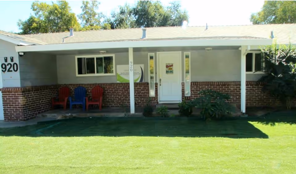 Front view of a single-story care home building with a covered porch. The porch has three Adirondack chairs, two red and one blue. The building has a brick lower half and gray siding upper half. The white front door is centered with two narrow windows on each side. The house number 920 is visible on the left side near the porch. There is a well-maintained green lawn in front and some bushes near the porch.
