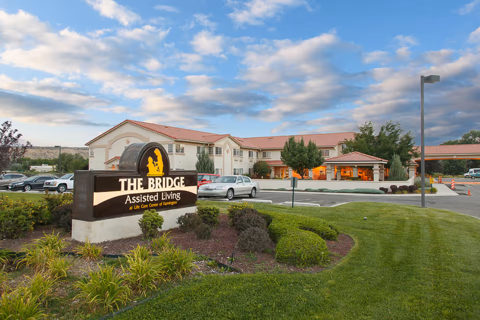 Exterior view of The Bridge Assisted Living facility at Life Care Center of Farmington, showing a large sign in the foreground with the facility name and a two-story building with a red-tiled roof in the background under a partly cloudy sky.