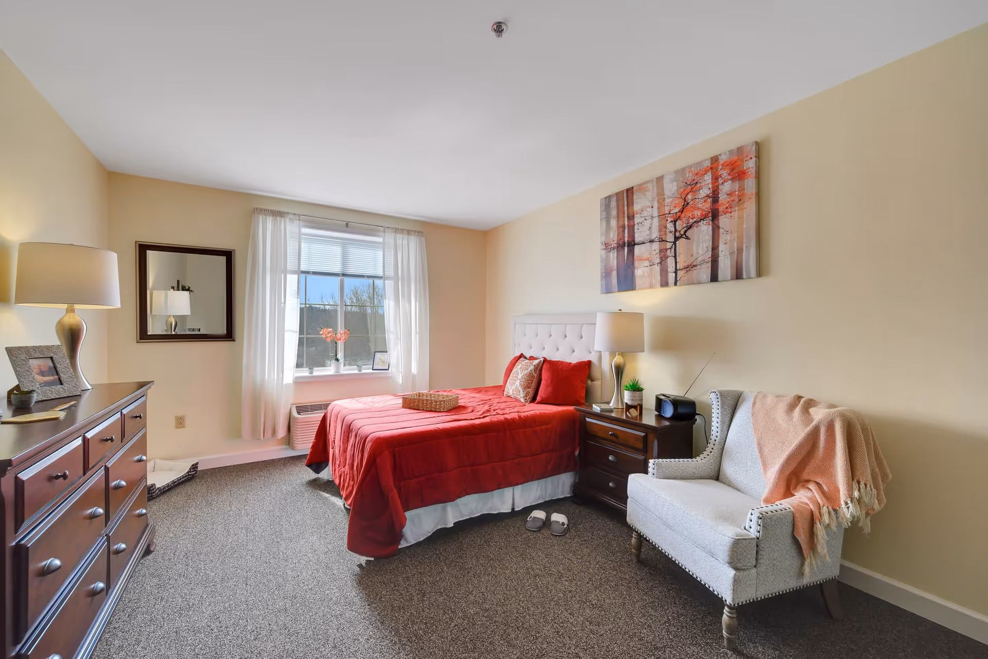 A cozy bedroom with a bed covered in a red comforter and matching pillows. There is a wooden dresser with a lamp and framed photo on the left side. A window with sheer white curtains lets in natural light. Next to the bed is a nightstand with a lamp, a small plant, and a radio. A light gray upholstered armchair with a pink throw blanket is positioned near the nightstand. A colorful painting of trees hangs on the wall above the bed.