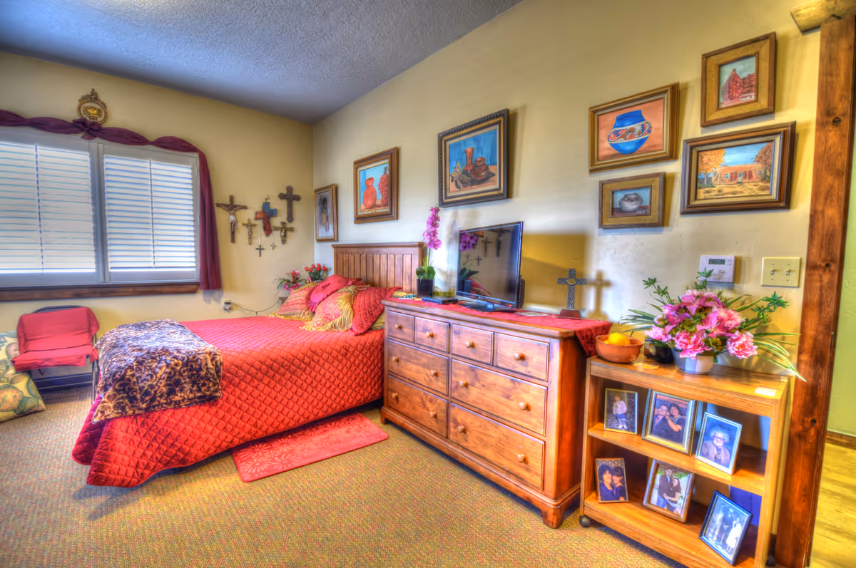 Bright, warmly decorated bedroom with a red quilted bed, wooden dresser and TV, wall art and framed photos.