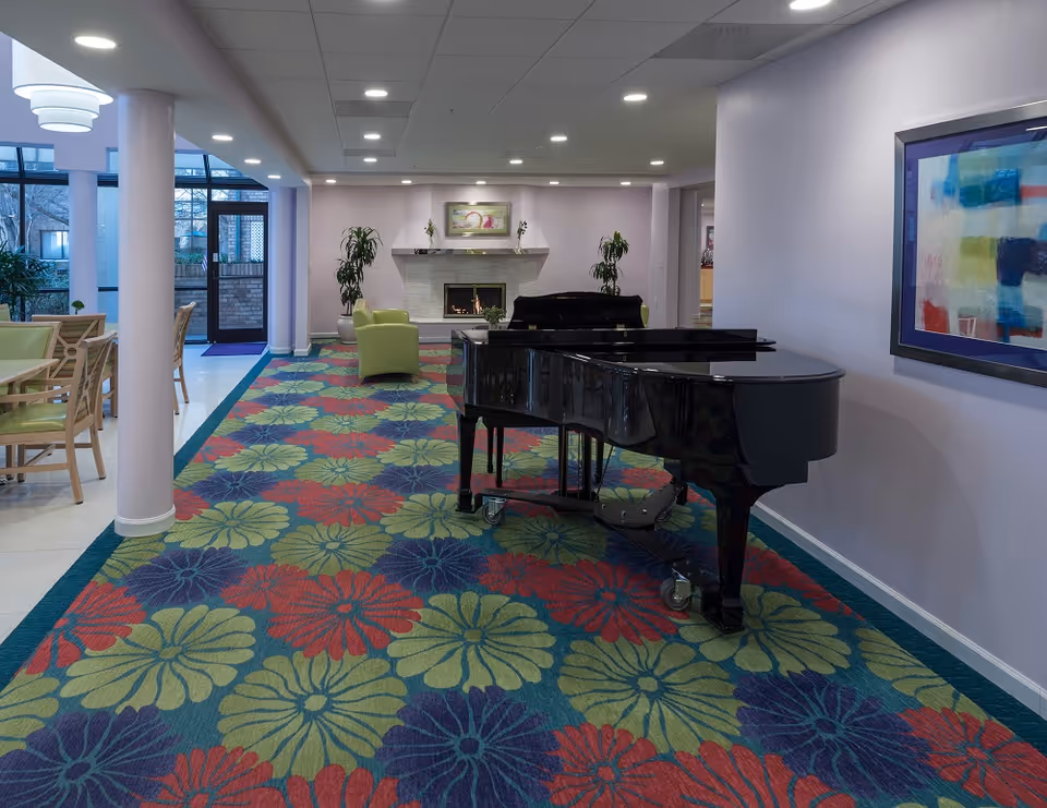 Interior of a senior living facility common area with a colorful floral carpet, a black grand piano on wheels, green armchairs, a fireplace with a painting above it, and large windows letting in natural light.