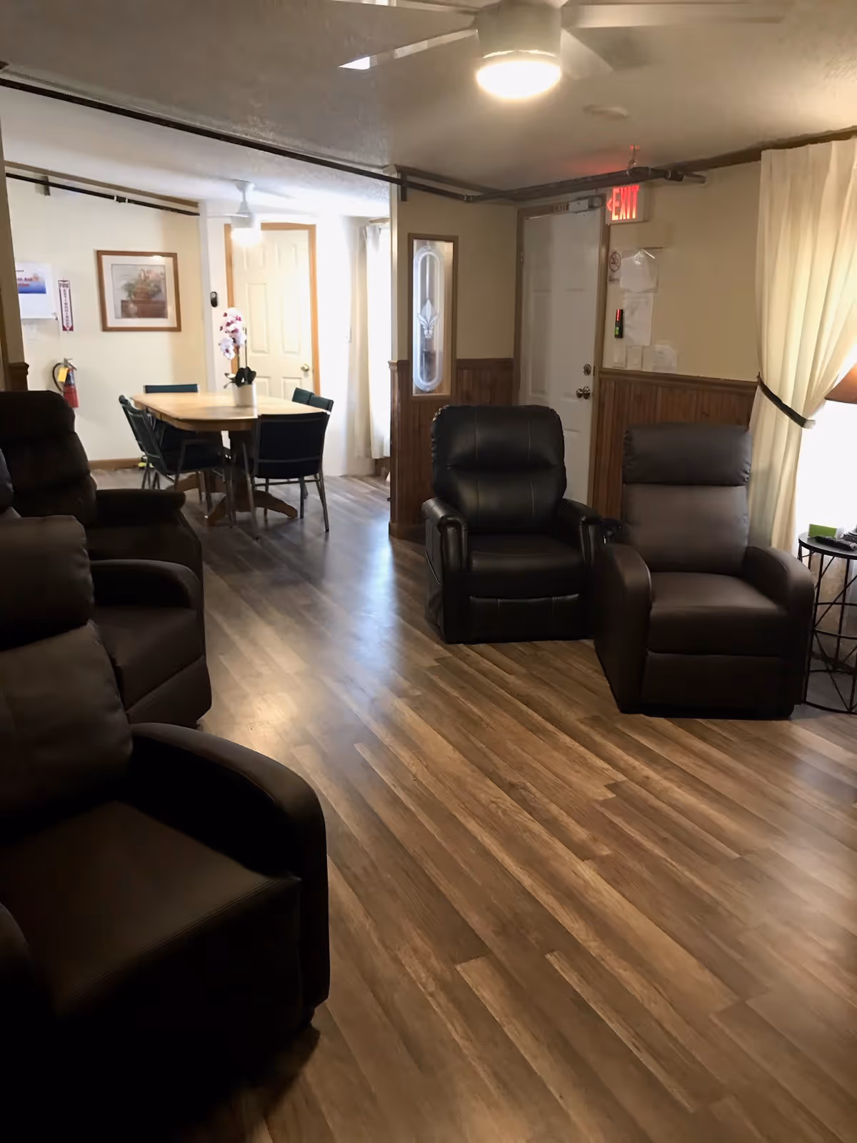 A senior living common room with several leather recliners, wood-look flooring, and a dining table in the background.
