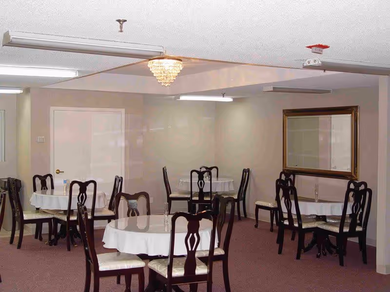 A dining room with several round tables covered with white tablecloths, each surrounded by dark wooden chairs with cushioned seats. The room has beige walls, a large mirror on one wall, and a chandelier hanging from the ceiling. The floor is carpeted in a muted red color.