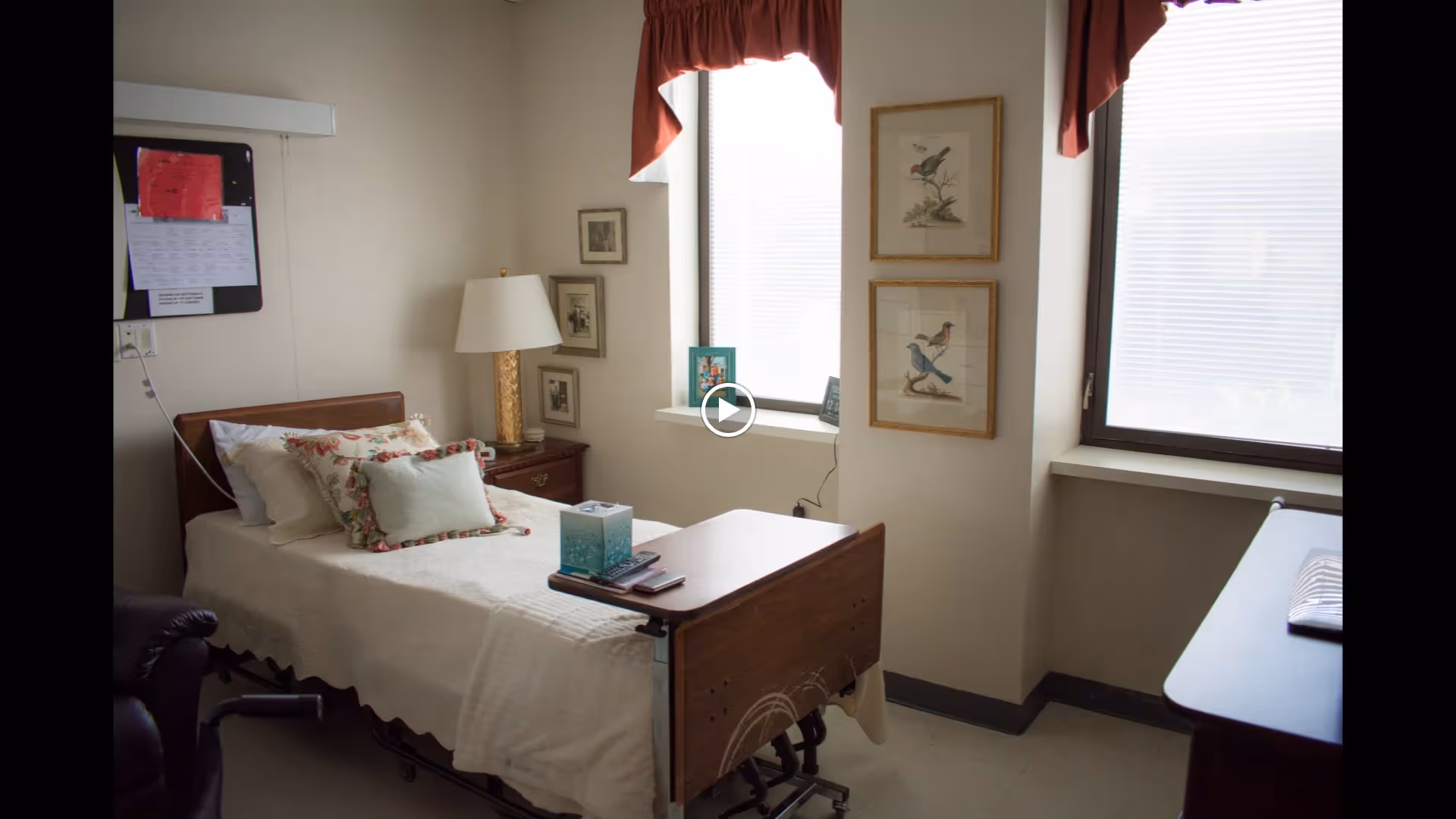 A small, neatly arranged bedroom in a healthcare facility with a single hospital-style bed covered in white bedding and decorative pillows. There is a wooden bedside table with a lamp and framed pictures on the wall. Two windows with red valances allow natural light into the room. A tissue box and remote controls are placed on a wooden overbed table.
