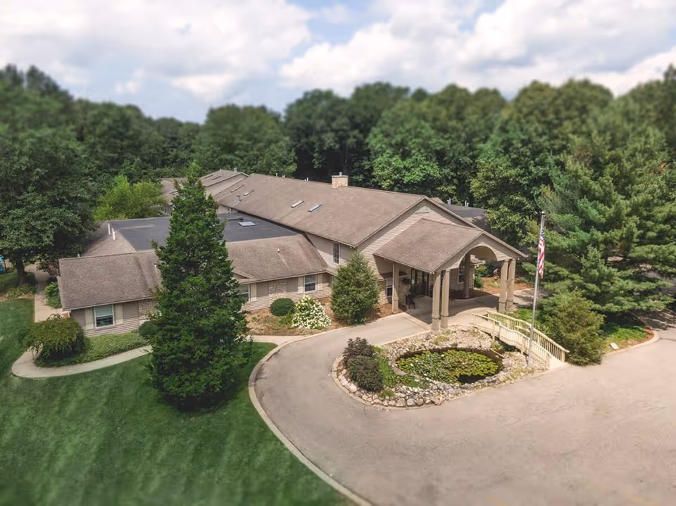 Aerial view of a single-story senior living facility named Golden Orchards surrounded by lush green trees and well-maintained landscaping. The building has a covered entrance with a small pond and a flagpole displaying the American flag in front. The sky is partly cloudy.