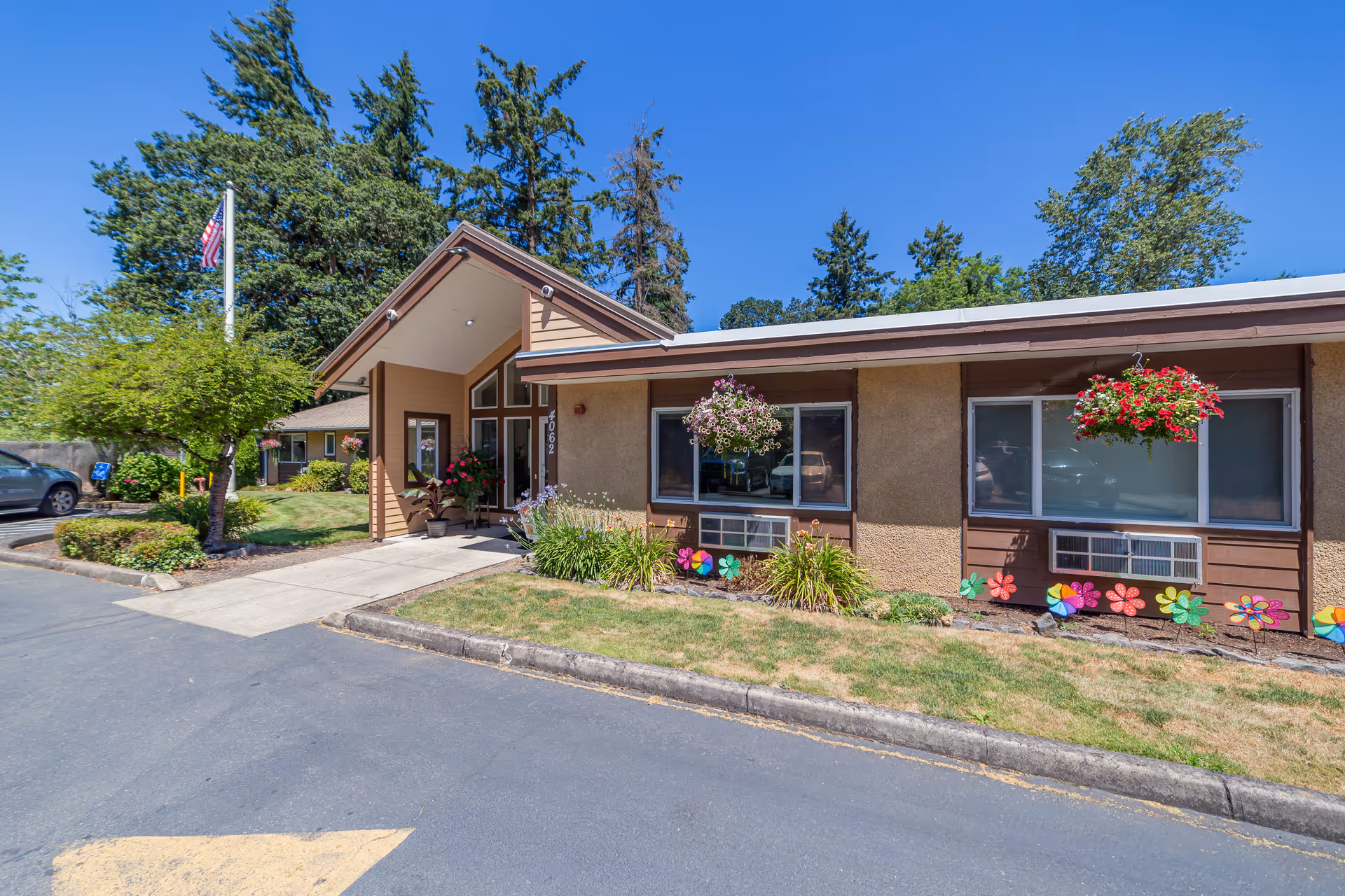 Exterior view of Keizer Nursing and Rehabilitation Center building on a sunny day, showing the entrance with a peaked roof, hanging flower baskets, colorful flower decorations near the windows, a small tree, and a flagpole with an American flag.