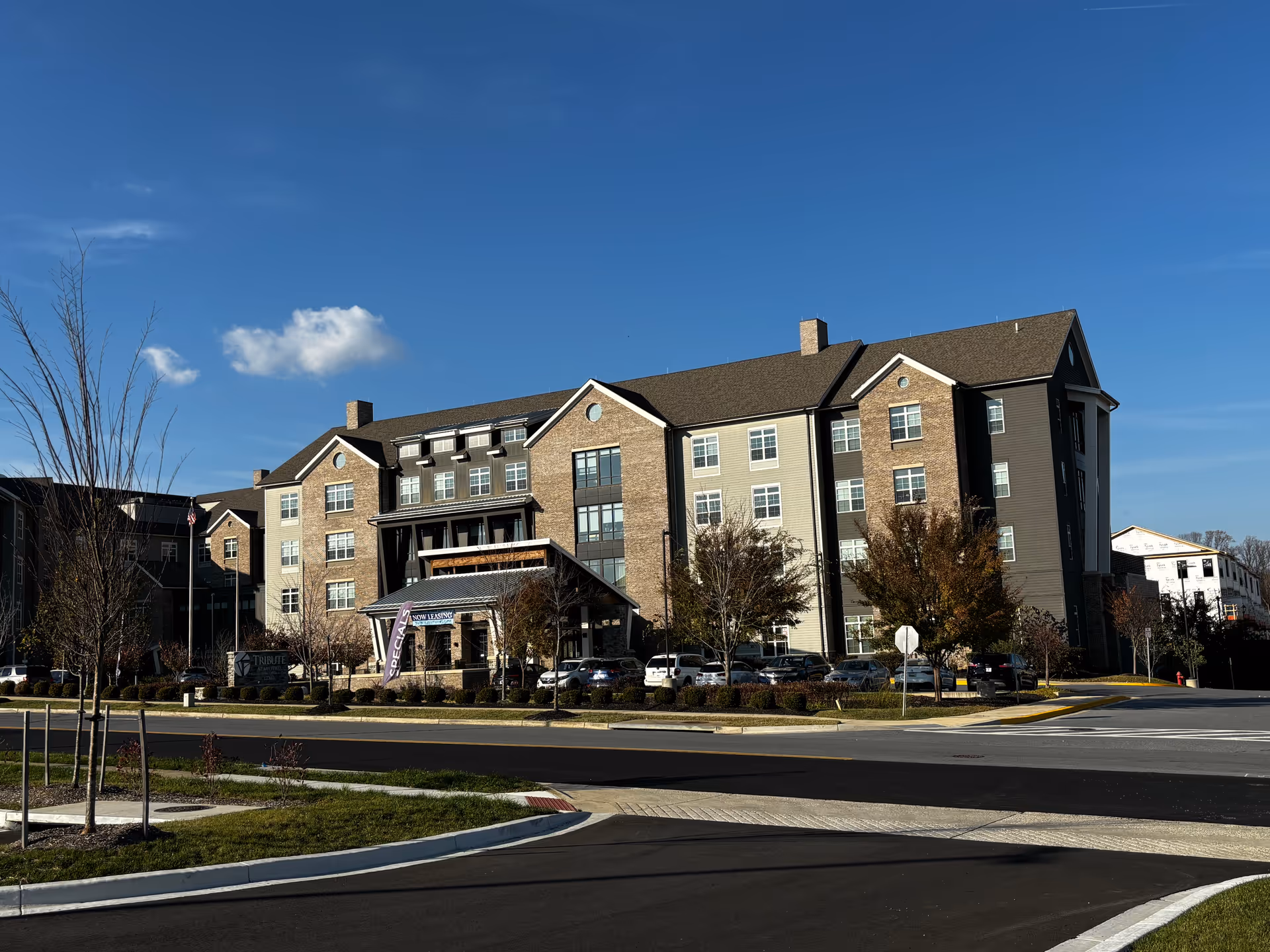 Large multi-story senior living building with a covered entrance, parking area and landscaped grounds under a clear blue sky.