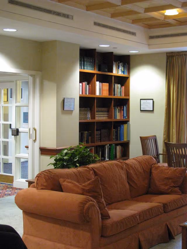 A cozy living room area with a brown sofa and matching cushions, a wooden bookshelf filled with books, a green potted plant, wooden chairs, and a framed certificate on the wall. The room has soft lighting and beige walls with a window covered by gold curtains.