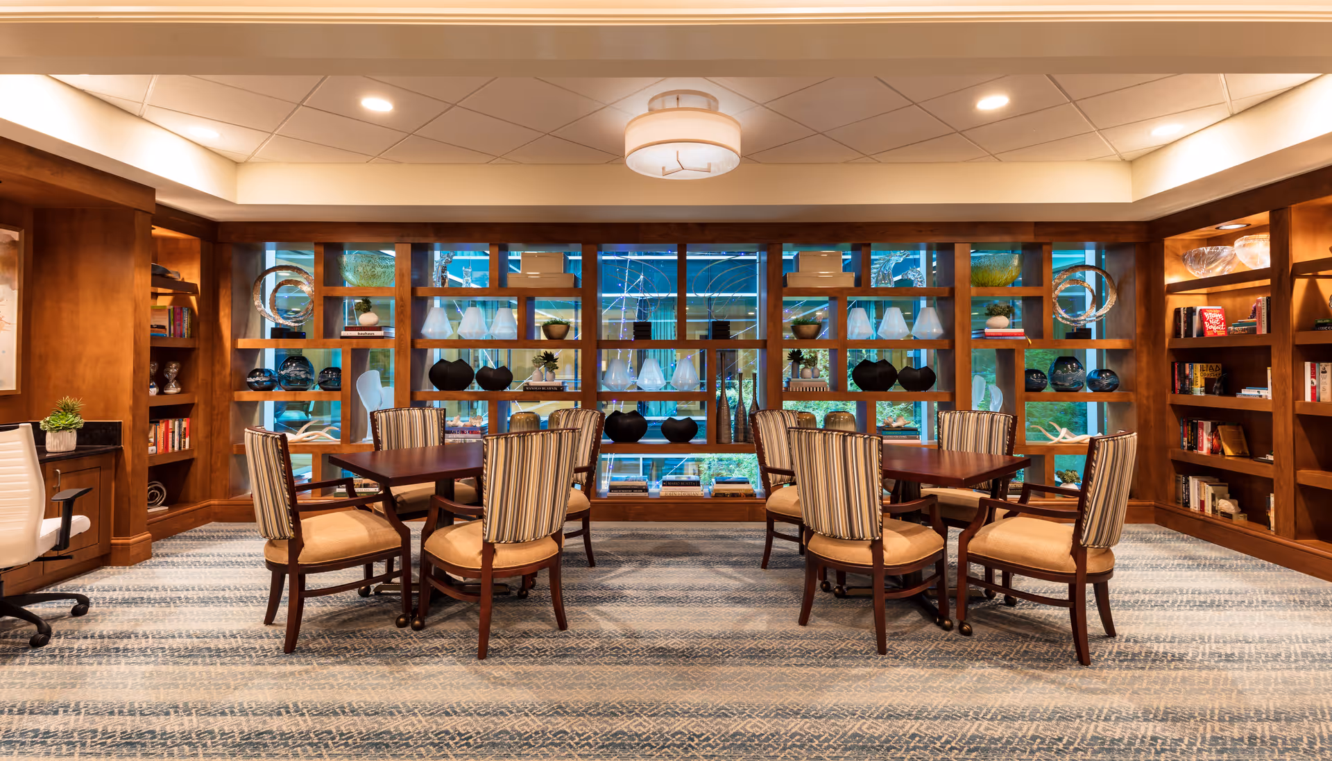 A well-lit interior room with two wooden tables, each surrounded by four cushioned chairs with striped backs. The back wall features a large wooden shelving unit with decorative items such as vases, books, and plants. The room has a patterned carpet and a modern ceiling light fixture.