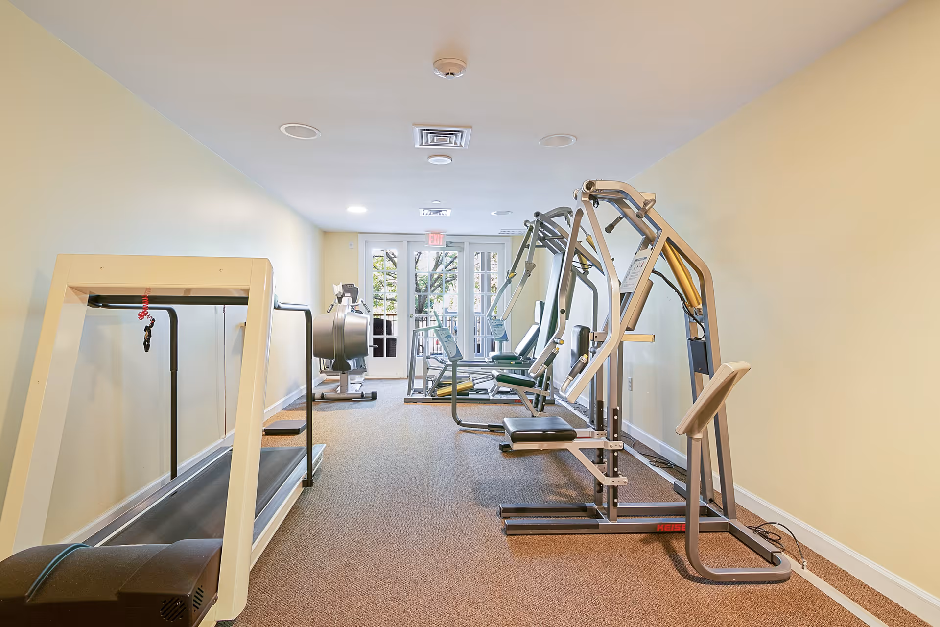 A small fitness room with various exercise equipment including a treadmill, weight machines, and a rowing machine. The room has beige walls, carpeted floor, and a glass door at the far end letting in natural light.