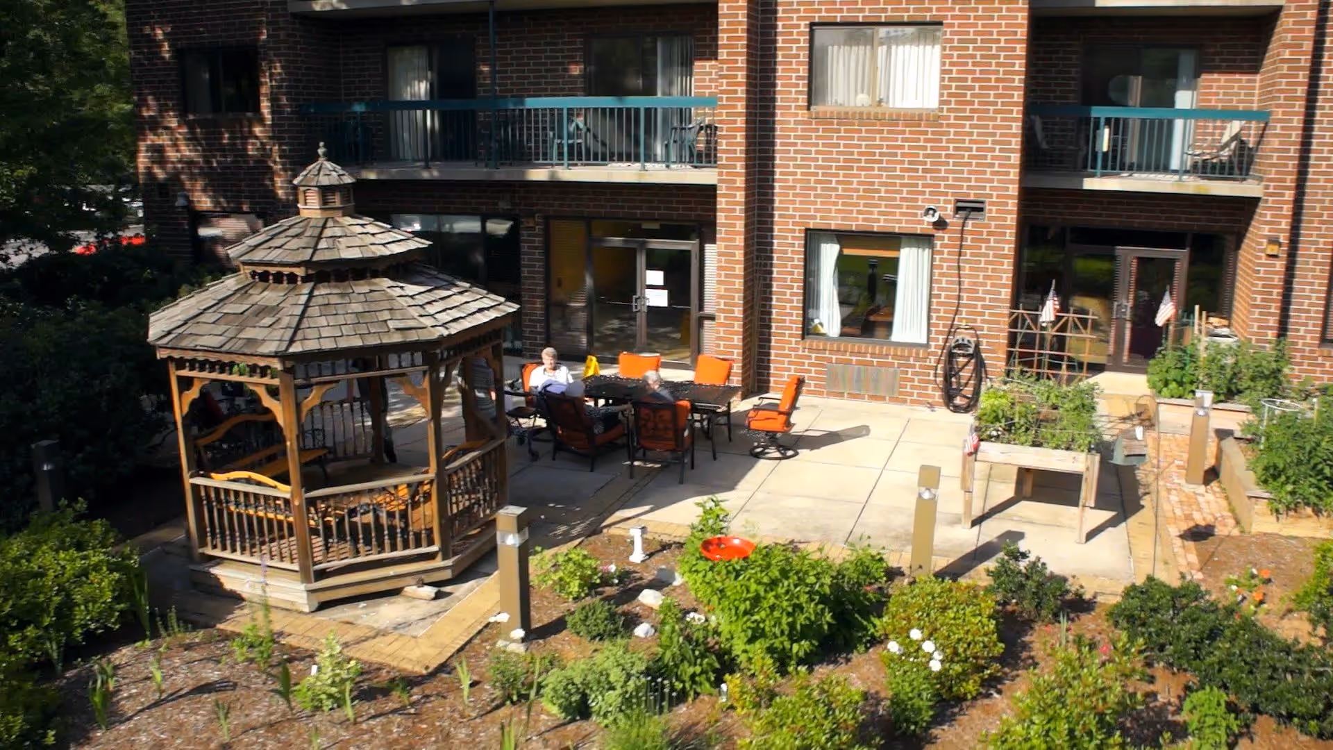 Outdoor patio area at The Woods at Cedar Run featuring a wooden gazebo, several orange cushioned chairs around a table, and a garden with various plants and flowers in front of a brick building with balconies and windows.