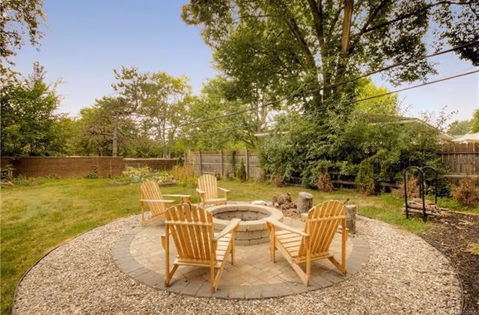 Outdoor seating area with four wooden Adirondack chairs arranged around a circular stone fire pit, surrounded by gravel and grass, with trees and a wooden fence in the background under a clear sky.