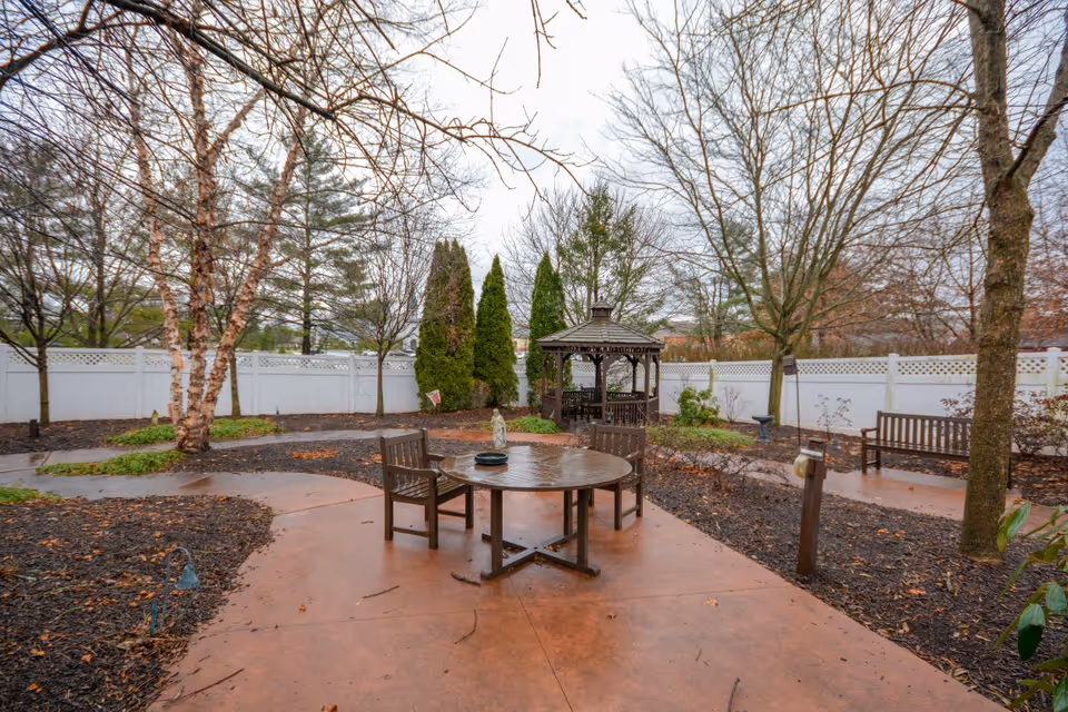 Outdoor courtyard with a round wooden table and chairs, a gazebo, benches, trees and a white fence.
