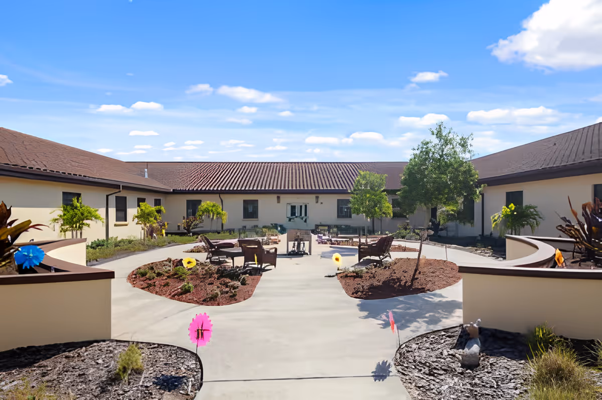 Outdoor courtyard area of a senior living facility with a clear blue sky, surrounded by single-story buildings with brown roofs. The courtyard features a paved walkway, small landscaped garden beds with young trees and plants, and several chairs arranged around a central fire pit or heater.