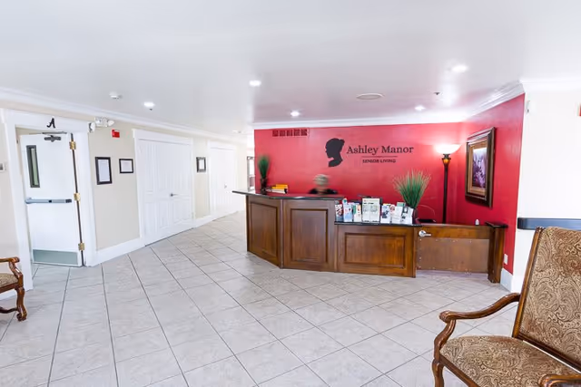 Reception area of Ashley Manor Senior Living with a wooden front desk against a red accent wall displaying the facility's name and logo. The floor is tiled, and there are chairs with patterned upholstery on the right side. The area is well-lit with ceiling lights and a floor lamp near the desk.