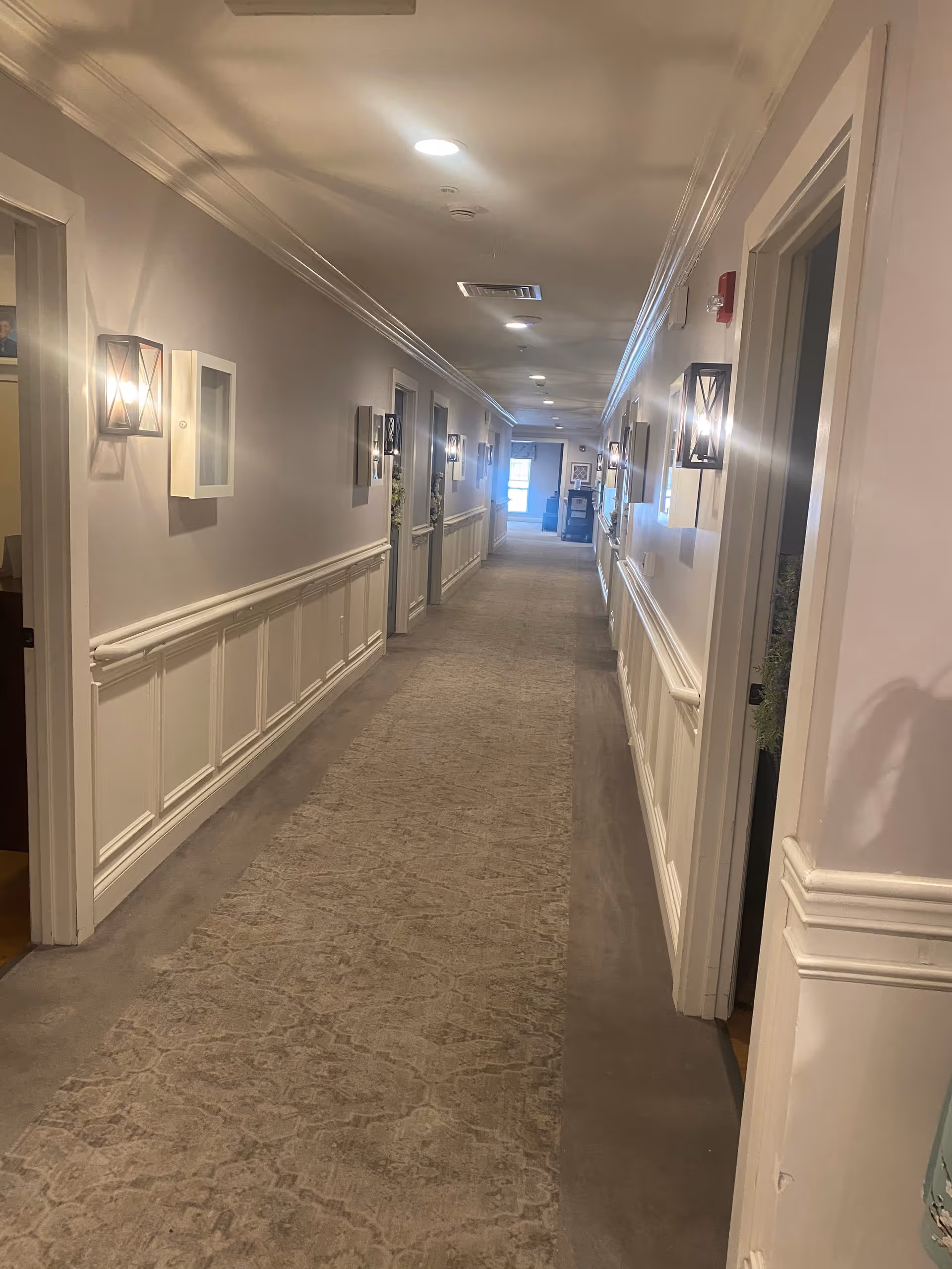 A long, well-lit hallway in a senior living facility with beige walls, white wainscoting, and a patterned carpet runner. Wall-mounted light fixtures line both sides of the corridor, and several doorways lead to rooms on either side. The hallway extends toward a bright window at the far end.