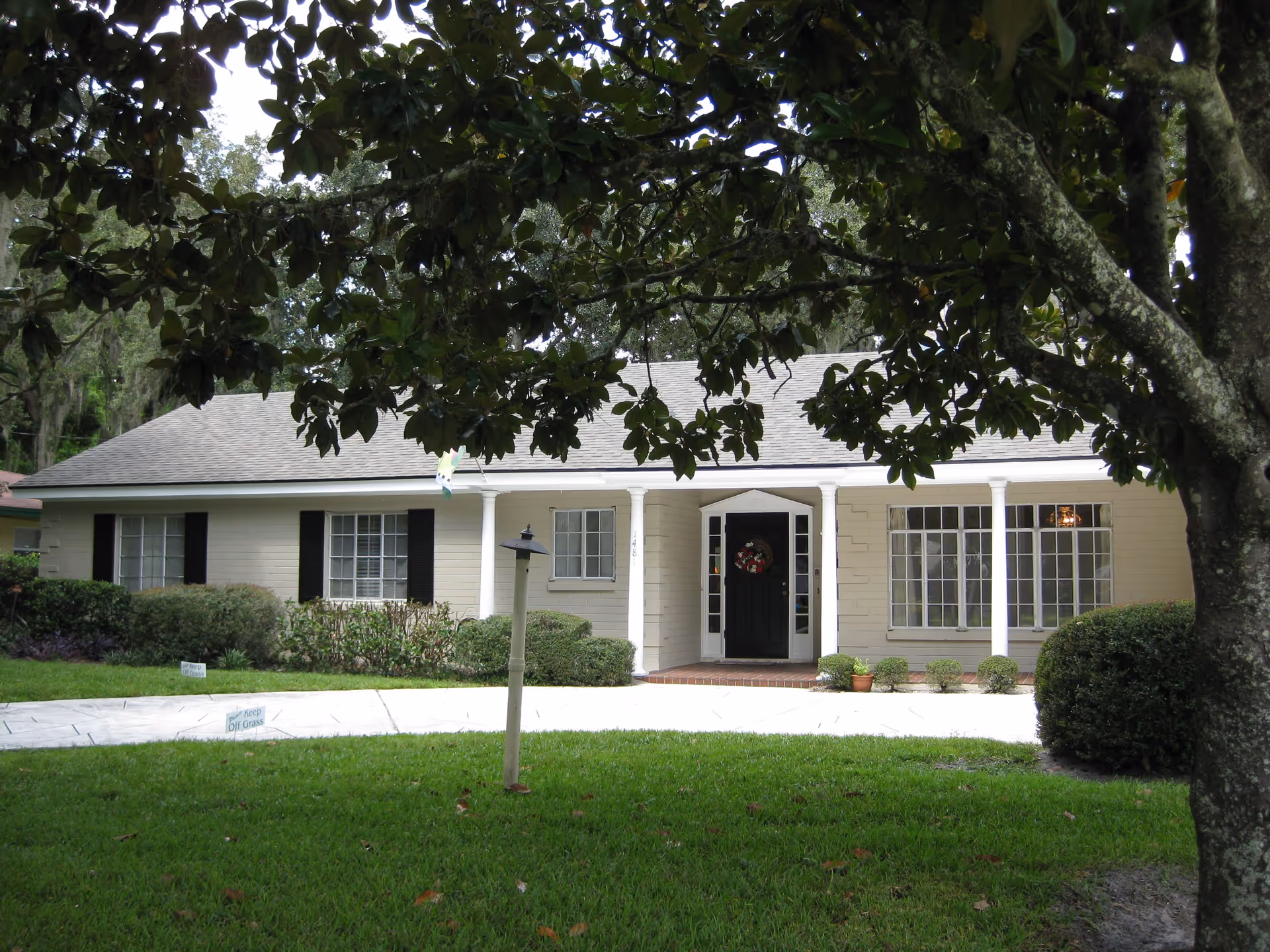Front exterior view of a single-story house with beige siding, black shutters, a black front door with a wreath, white columns on the porch, and a well-maintained green lawn with bushes and a tree in the foreground.