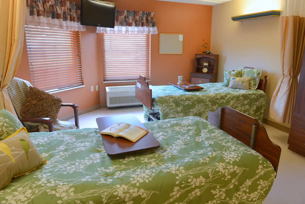 A bright bedroom with two single beds covered in green floral bedspreads. Each bed has a wooden headboard and a tray on top, one with an open book and the other with a cup. There is a cushioned armchair with a brown pillow near two windows with wooden blinds and floral valances. A small wooden nightstand with a clock and decorative items is against the wall, and a flat-screen TV is mounted above the windows.