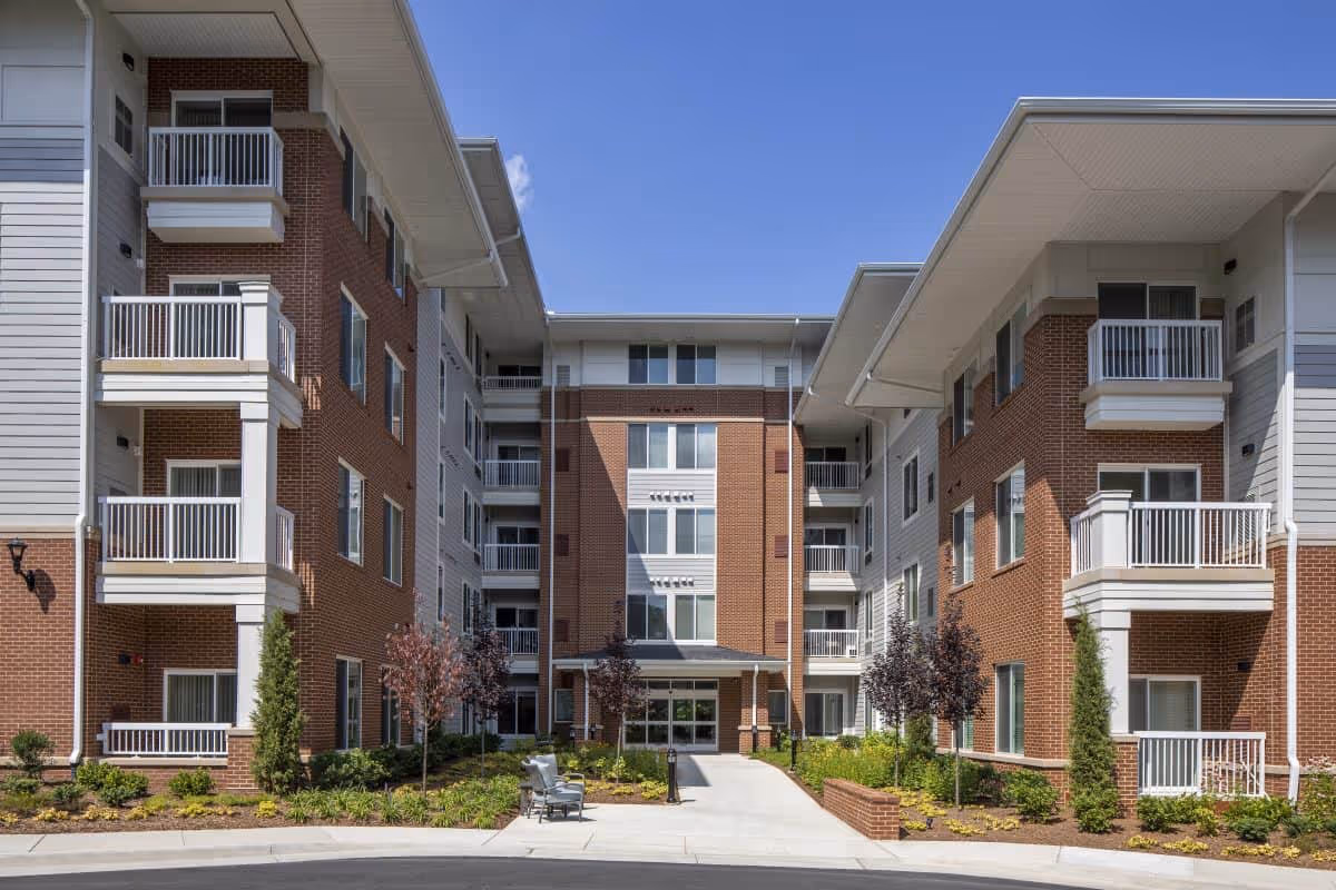 Front entrance of a multi-story brick and siding senior living facility with balconies and a landscaped walkway.