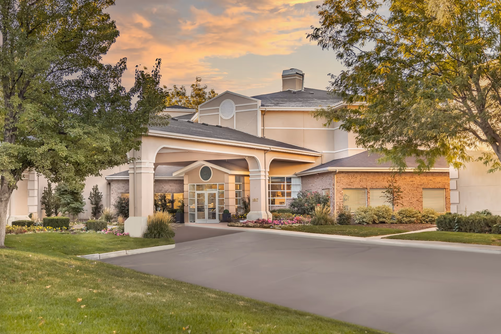Exterior view of Legacy House of South Jordan building at sunset, showing the entrance with a covered driveway, surrounded by trees, shrubs, and well-maintained landscaping.