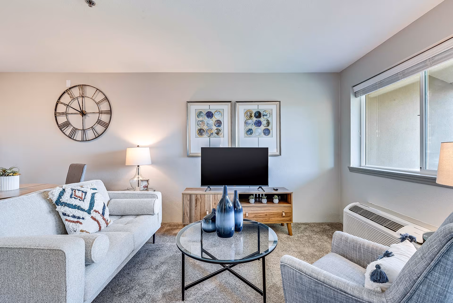 Bright living room with a sofa and armchair around a glass coffee table, a TV on a wooden console, wall clock, and window.