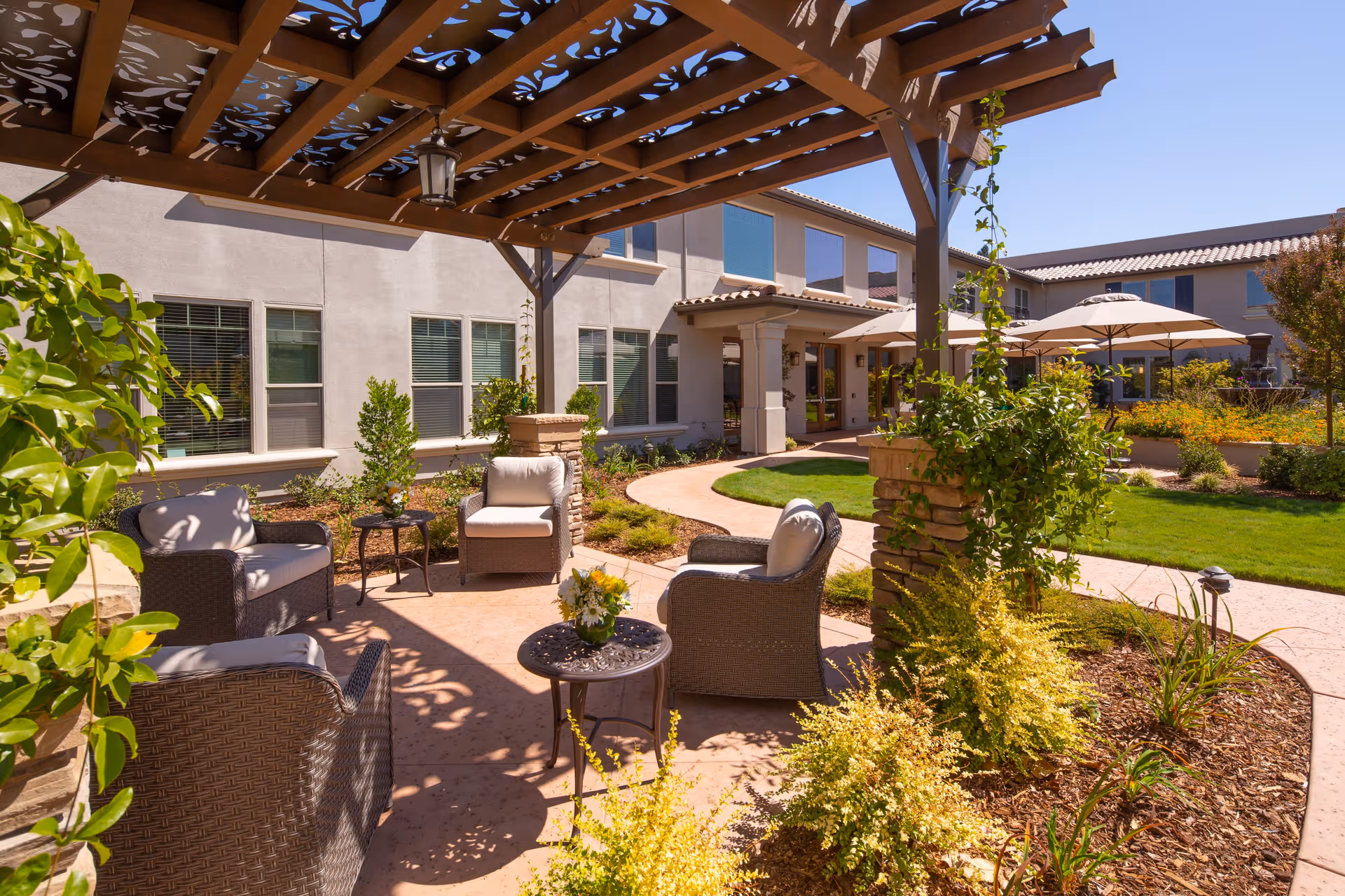 Outdoor seating area at Oakmont of Fair Oaks featuring cushioned wicker chairs and small tables under a wooden pergola with climbing plants. The area is surrounded by landscaped garden beds and a curved walkway, with the building visible in the background under a clear blue sky.