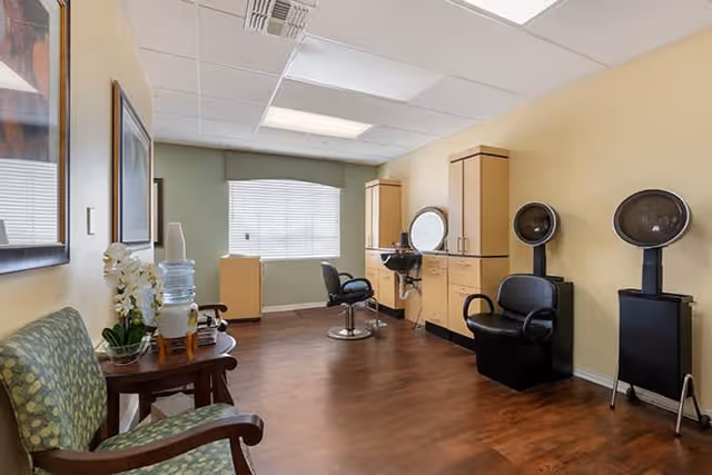 Interior view of a salon area in a senior living facility featuring two black salon chairs with hair dryers, a styling station with a round mirror, a window with blinds, a green patterned armchair, a small table with a water dispenser and a plant, and light yellow walls with framed artwork.