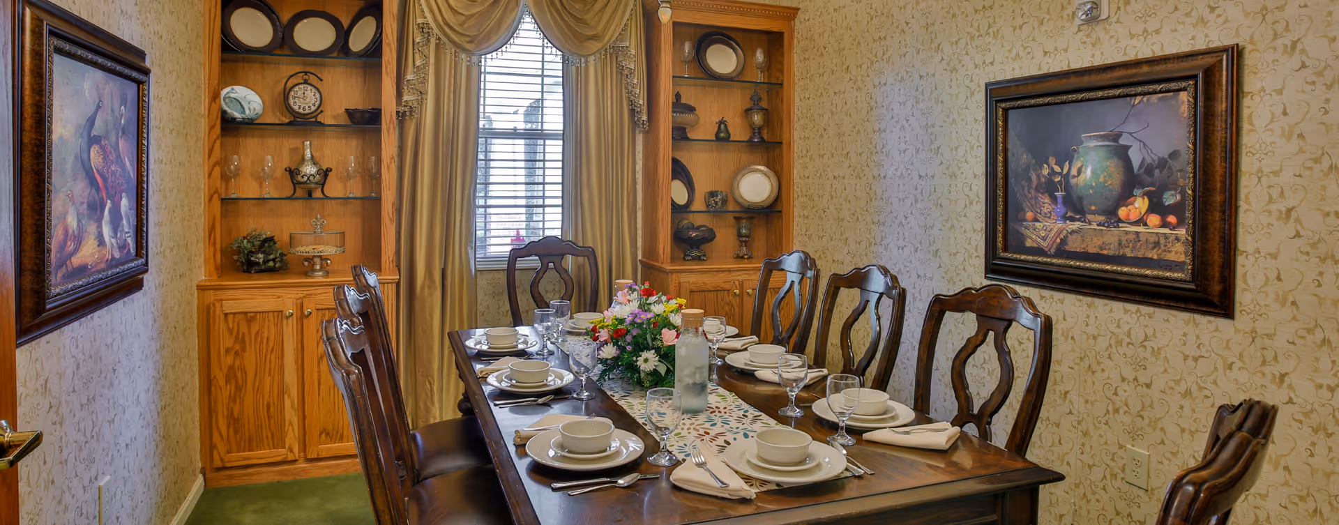 A traditional dining room with a wooden dining table set for eight people. The table is adorned with white plates, bowls, glasses, silverware, and beige napkins, along with a floral centerpiece and a water bottle. The room features patterned wallpaper, two wooden display cabinets with decorative plates and glassware, a window with beige curtains, and framed paintings on the walls.